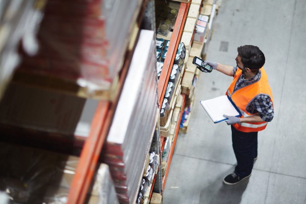Warehouse worker using an RF scanner to check inventory on warehouse shelving while holding a clipboard in a distribution centre.