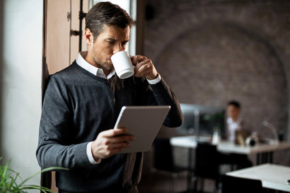 male-entrepreneur-surfing-net-digital-tablet-while-drinking-coffee-by-window-office