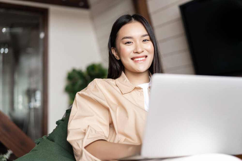 technology and IT - smiling-asian-woman-using-laptop-computer-home-looking-happy-camera