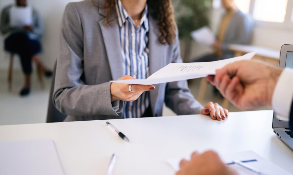 In the office young Caucasian female candidate, giving her CV or application form to male recruiter during a job interview