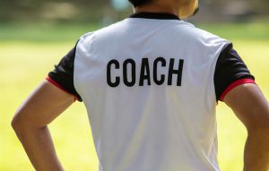 Person standing with their hands on their hips facing away from the camera, and they have the word Coach printed across the back of their shirt
