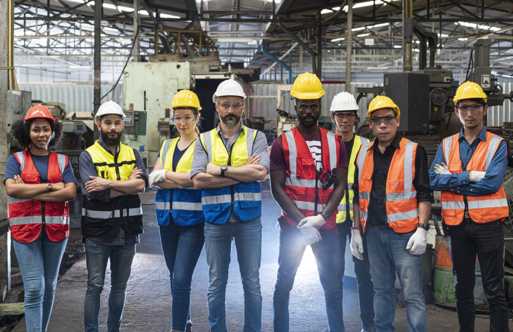 Group of diverse trade and engineering workers wearing safety gear and PPE, standing together confidently in an industrial workshop.