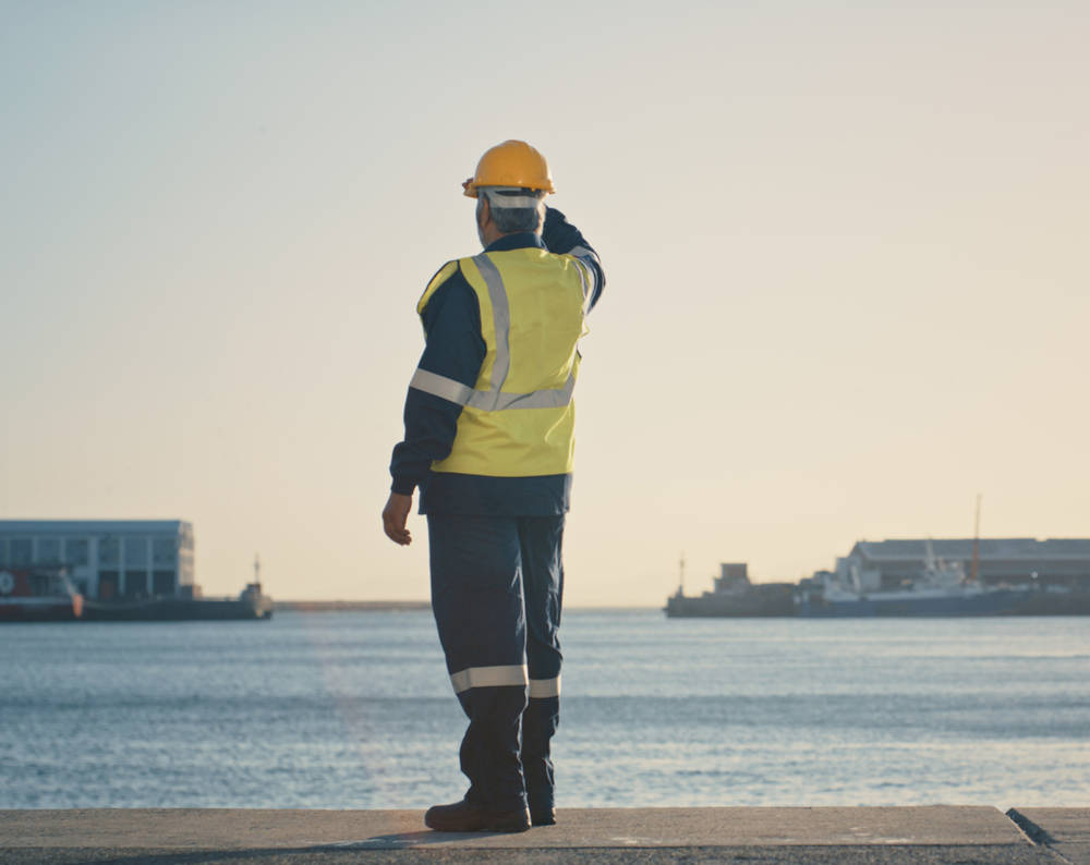 Shipping, freight and logistics with a supervisor standing on the dock in a harbor, looking at the view and waiting for a delivery or shipment. Safety and control in the import and export industry