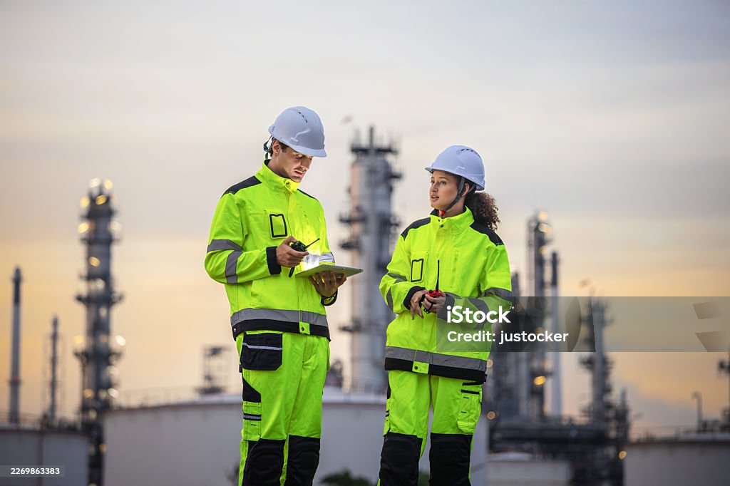 Industrial engineers in safety uniforms using digital tablet and radio at oil refinery during sunset, Maintenance crew in reflective workwear and hard hats discussing operations at fuel power station